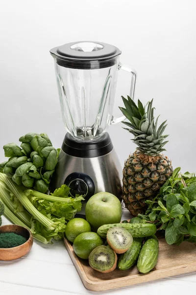 Licuadora cerca de verduras verdes, frescas y frutas crudas orgánicas en blanco - foto de stock