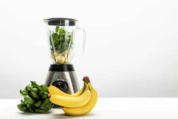 Green fresh spinach leaves and yellow ripe bananas near blender on white — Stock Photo