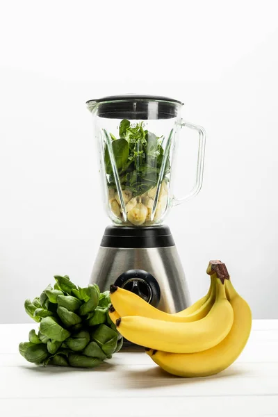 Green spinach leaves and yellow ripe bananas near blender on white — Stock Photo