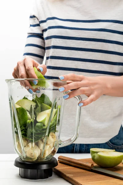 Vista recortada de la mujer poniendo manzana verde en licuadora con plátanos y hojas de espinaca en blanco - foto de stock