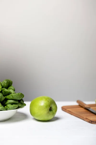 Assiette avec des feuilles d'épinards près de pomme verte et planche à découper sur gris — Photo de stock