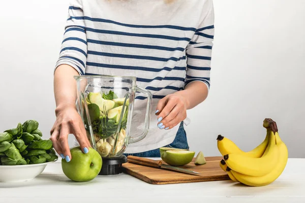 Vista ritagliata di donna che prende mela biologica vicino frullatore con ingredienti su bianco — Foto stock