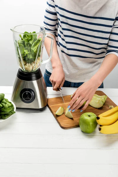 Cropped view of woman cutting organic apple near blender with ingredients on white — Stock Photo