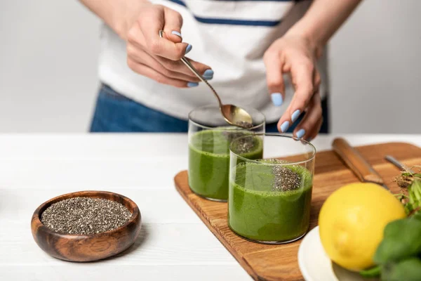 Cropped view of woman holding spoon with chia seeds near glasses of green smoothie on grey — Stock Photo