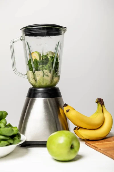 Зеленое вкусное яблоко возле листьев шпината, бананов и блендера на сером — стоковое фото