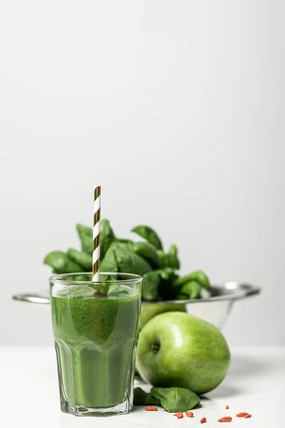 Foco selectivo de batido verde en vidrio con paja cerca de hojas de espinacas frescas y manzana en gris - foto de stock