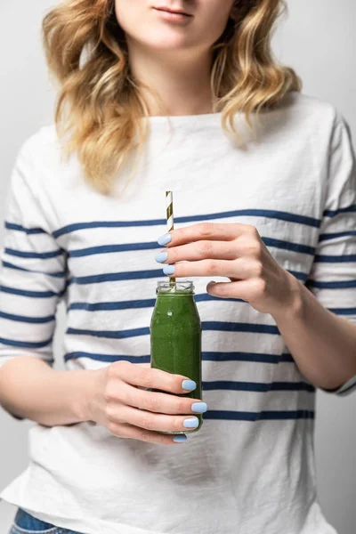 Обрезанный вид женщины, держащей солому и стеклянную бутылку с вкусным зеленым смузи на белом — стоковое фото
