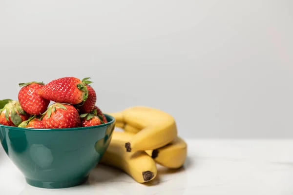 Fragole dolci e saporite in ciotola vicino a banane gialle su grigio — Foto stock