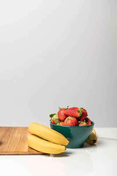 Fresas dulces en tazón cerca de plátanos amarillos y tabla de cortar en gris - foto de stock