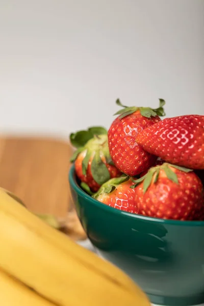 Selective focus of red strawberries in bowl near yellow bananas on grey — Stock Photo