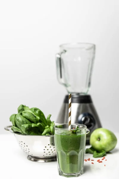 Вибірковий фокус зеленої смузі в склянці з соломою біля свіжого листя шпинату, смачне яблуко і блендер на білому — стокове фото