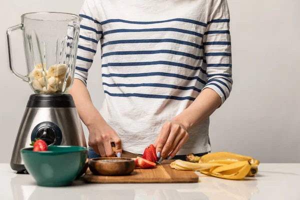 Vista ritagliata di donna che taglia fragole dolci vicino a banane e frullatore su bianco — Foto stock