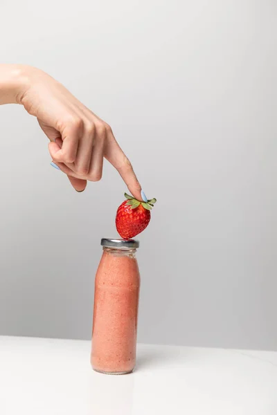 Vista cortada de mulher segurando morango maduro perto de garrafa de vidro com saboroso smoothie rosa no cinza — Fotografia de Stock