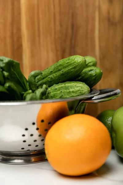 Selective focus of green cucumbers in colander near orange and wooden cutting board — Stock Photo