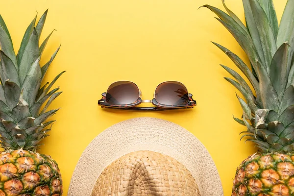 Вид зверху на солом'яний капелюх і сонцезахисні окуляри біля ананасів на жовтому фоні — стокове фото