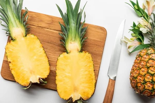 Top view of cut ripe yellow pineapple on wooden chopping board near knife and Alstroemeria flowers on white background — Stock Photo