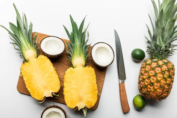 Draufsicht auf geschnittene reife gelbe Ananas auf hölzernem Schneidebrett in der Nähe von Kokosnusshälften, Limetten und Messer auf weißem Hintergrund — Stockfoto