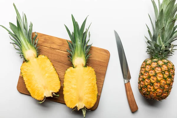 Draufsicht auf geschnittene reife gelbe Ananas auf hölzernem Schneidebrett neben ganzen Früchten und Messer auf weißem Hintergrund — Stockfoto