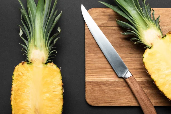 Top view of cut ripe pineapple on wooden chopping board with knife on black background — Stock Photo