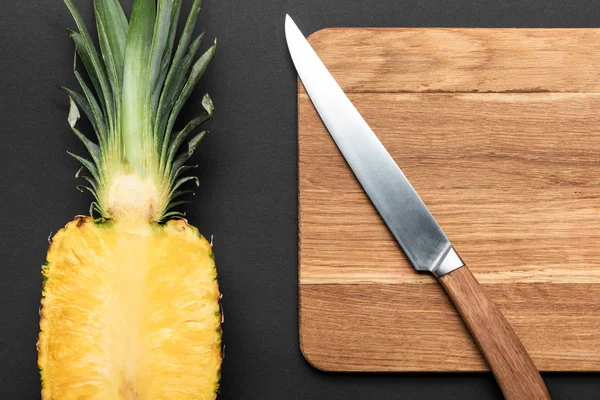 Top view of ripe pineapple half on wooden chopping board with knife on black background — Stock Photo