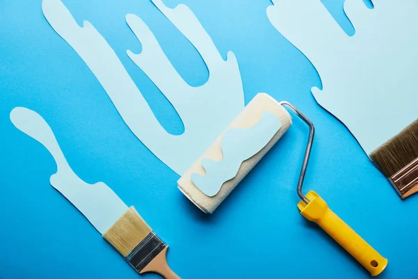 Vista superior do rolo e escovas com tinta de corte de papel gotejamento no fundo azul brilhante — Fotografia de Stock
