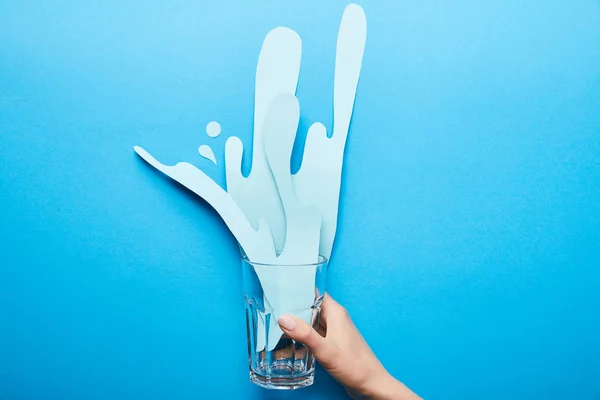Cropped view of woman holding glass with paper cut splash on blue background — Stock Photo