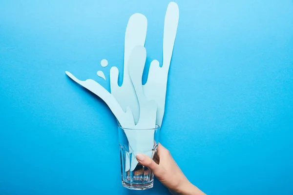 Vista recortada de la mujer sosteniendo vidrio con salpicadura de papel cortado sobre fondo azul - foto de stock