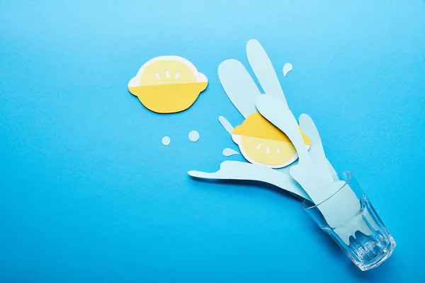Top view of glass with paper cut water splash and lemons on blue background with copy space — Stock Photo