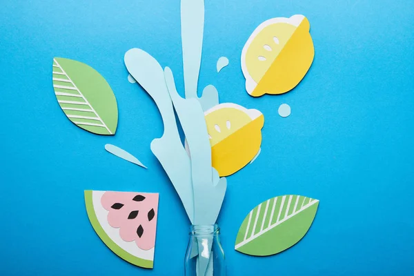 Top view of bottle with paper cut water splash, lemons, leaves and watermelon on blue background — Stock Photo