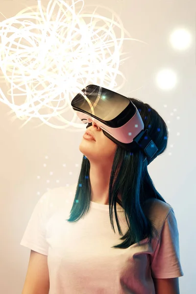 Молодая женщина в наушниках виртуальной реальности смотрит на светящиеся абстрактные иллюстрации на бежевом фоне — стоковое фото