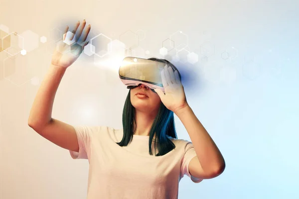 Young woman in virtual reality headset pointing with hands at glowing abstract  illustration on beige and blue background — Stock Photo