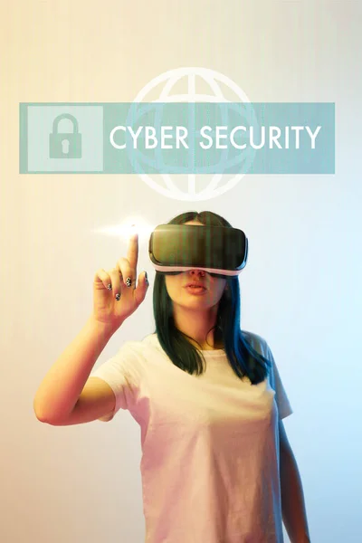 Young woman in vr headset pointing with finger at cyber security illustration on beige and blue background — Stock Photo