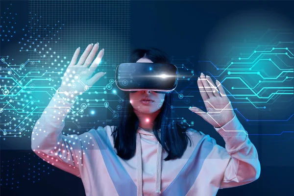 Mujer joven en auriculares de realidad virtual gesticulando entre la ilustración cibernética brillante sobre fondo oscuro - foto de stock
