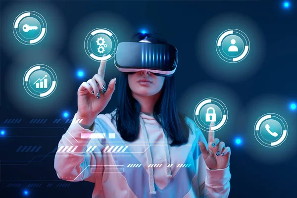 Young woman in virtual reality headset pointing with fingers at glowing cyber icons on dark background — Stock Photo