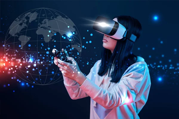 KYIV, UKRAINE - APRIL 5, 2019: Young woman in virtual reality headset using joystick on dark background with globe illustration — Stock Photo
