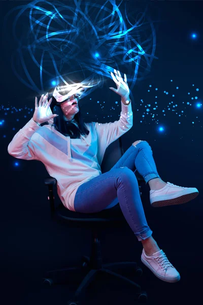 Young shocked woman in virtual reality headset sitting on chair and gesturing among glowing data illustration on dark background — Stock Photo