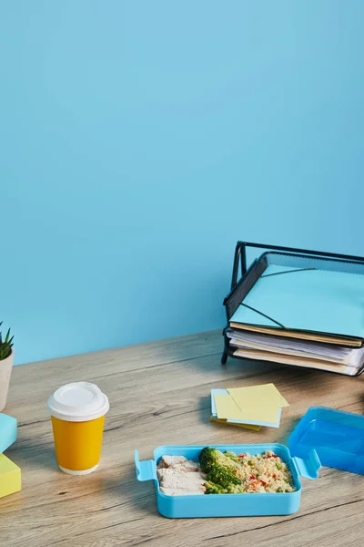 Scatola da pranzo con risotto, pollo e broccoli sul posto di lavoro con carte su tavolo di legno su sfondo blu — Foto stock