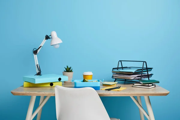 Рабочее место с бумагами и обеденной коробкой с кофе в пластиковой чашке на деревянном столе на синем фоне — стоковое фото
