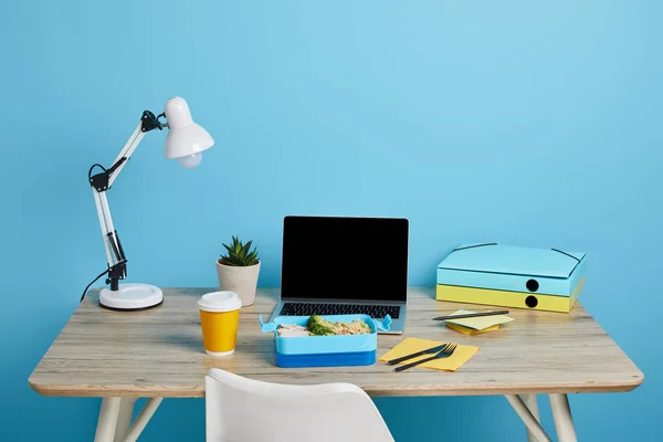 Workplace with laptop and lunch box on wooden table on blue background, illustrative editorial — Stock Photo