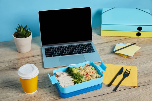 Обідня коробка з рисом, куркою та брокколі на робочому місці з ноутбуком на дерев'яному столі на синьому фоні, ілюстративна редакція — стокове фото