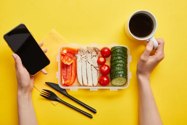 Обрізаний вид жінки зі смачним обідом, що тримає смартфон і чашку кави на жовтому фоні — стокове фото