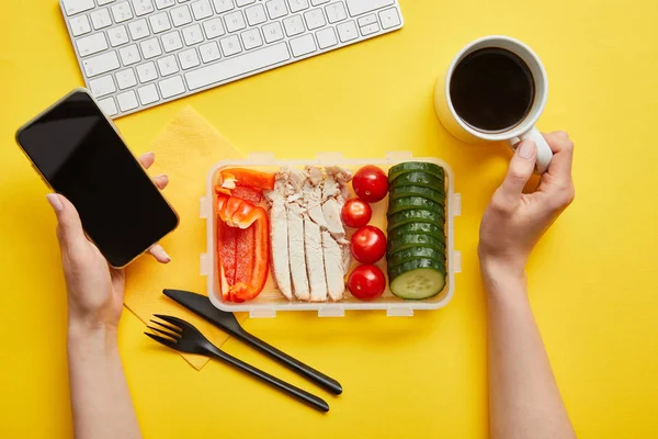 Cropped view of woman with healthy delicious lunch holding smartphone and cup of coffee on yellow background — Stock Photo