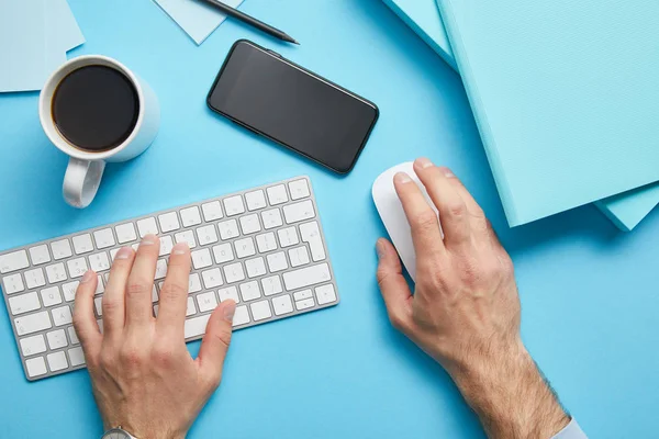 Vista recortada del hombre utilizando el teclado de la computadora y el ratón de la computadora en el lugar de trabajo con papeles, teléfono inteligente y taza de café sobre fondo azul - foto de stock