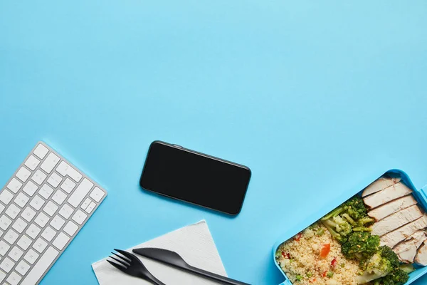 Vista superior do local de trabalho com smartphone, teclado de computador e lancheira com risoto, frango e brócolis em fundo azul — Fotografia de Stock