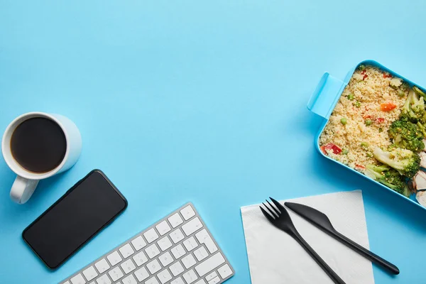 Top view of workplace with digital devices, cup of coffee and lunch box with risotto, chicken and broccoli on blue background — Stock Photo