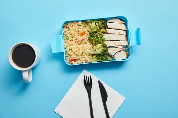 Top view of cup of coffee, disposable fork with knife and lunch box with risotto, chicken and broccoli on blue background — Stock Photo