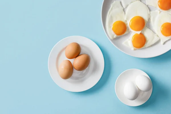 Вид сверху на свежие вареные и жареные яйца на белых тарелках на синем фоне — стоковое фото