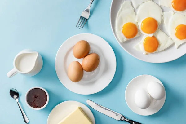 Top view of boiled and fried eggs, butter, jam on white plates, milk, fork, spoon and knife on blue background — Stock Photo