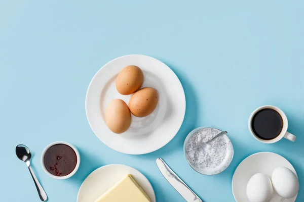 Vista superior de huevos recién cocidos, mantequilla, mermelada en platos blancos, yogur con semillas de chía, café, cuchara y cuchillo sobre fondo azul - foto de stock