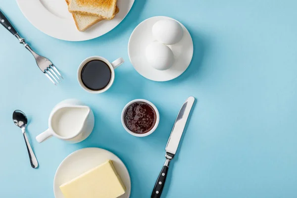 Vue de dessus des œufs frais, confiture, café, couverts, beurre et deux toasts sur des assiettes blanches sur fond bleu — Photo de stock